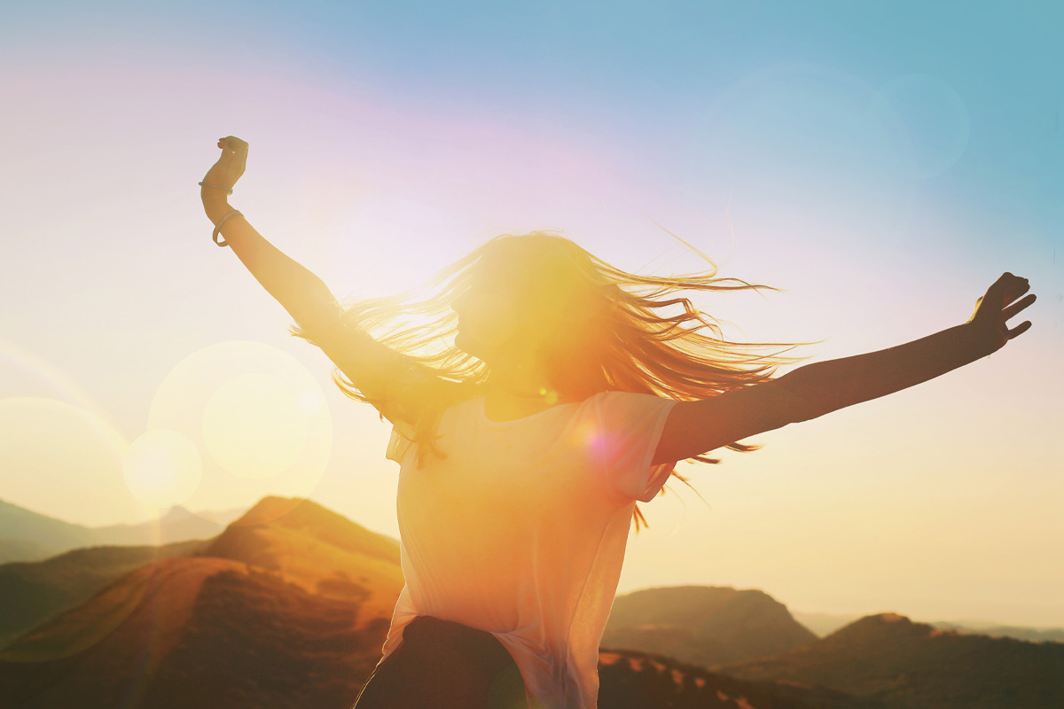 woman jumping up in the air looking happy with the sun shining behind her